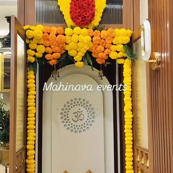 A white pooja unit with an Om panel in a flat, framed in long strings of yellow marigold, with a marigold swag across the top and a large red rose teardrop set in a yellow marigold header above it