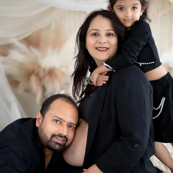 A pregnant woman in a black blazer on a bed set with white drapes and pampas, her husband resting his head on her bump and a young girl in black leaning on her back