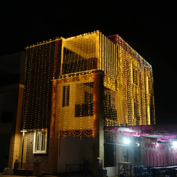 A three-storey house at night covered from roof to ground in strings of warm golden lights, with a marigold swag over the entrance and a pink-draped dining tent at the lower right