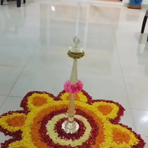 A round rangoli of yellow, orange and dark red petals shaped like a flower, with a tall brass lamp in the middle, on the tiled floor of an empty flat