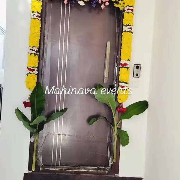 A flat's front door framed in yellow marigold and white flower strings, with a header of pink roses and purple and white flowers, two young banana plants in front and a small white rangoli at the step