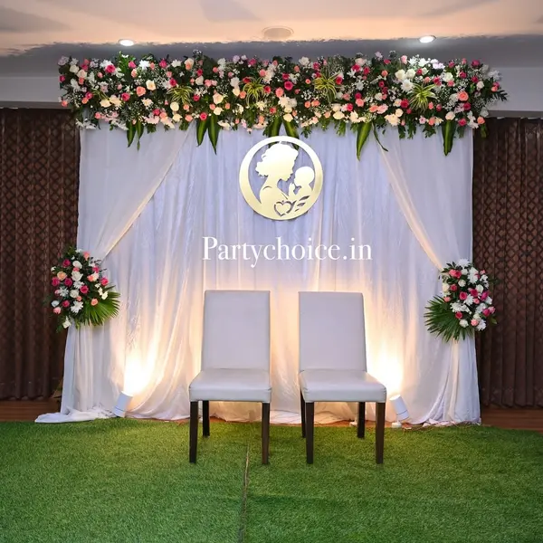 A baby shower backdrop: white drapes under a header of pink, white and peach roses and leaves, a gold mother-and-baby medallion, two flower stands and two white chairs, on a grass carpet