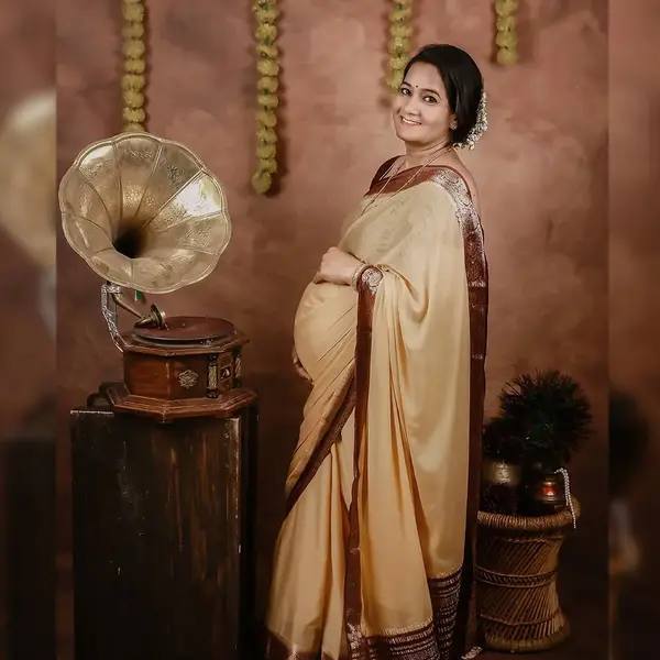 A pregnant woman in a beige saree with a maroon border stands beside a brass gramophone on a wooden stand, with marigold strings on a warm brown wall