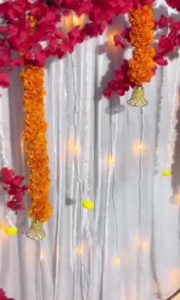 A white shimmer curtain hung with long orange marigold strings ending in gold bells, under branches of red blossoms, with warm lights behind