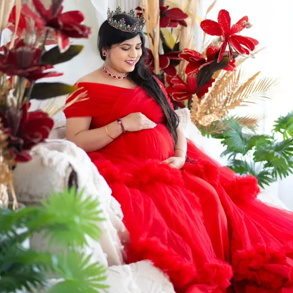 A pregnant woman in a red ruffled gown and a crown sits on a studio couch among dried flowers, one hand on her bump