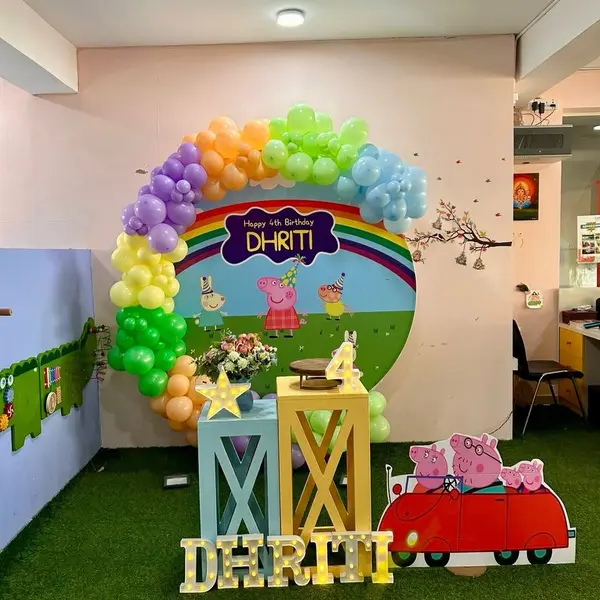 A Peppa Pig fourth birthday in a play-school room: a round print reading Happy 4th Birthday Dhriti in a rainbow garland, two painted plinths, a lit 4 and star, a cutout of Peppa's family in a red car, the name in lit letters and a grass carpet