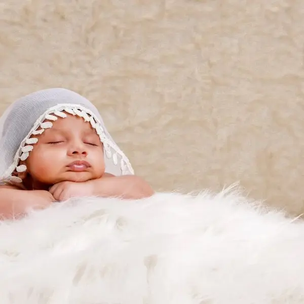 A newborn asleep with the chin on crossed hands under a white lace veil edged with pom-poms, on white fur against a cream wall