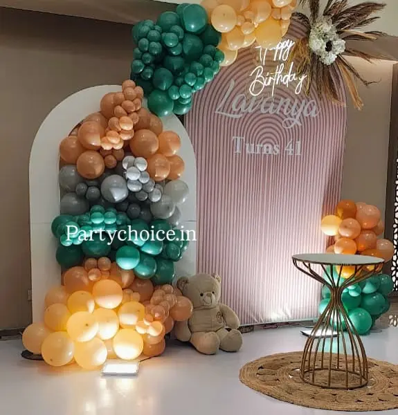 A 41st birthday: a white arched panel and a blush striped panel reading Lavanya Turns 41, a peach, teal and grey garland, a pampas spray, a Happy Birthday neon sign, a teddy, a jute rug and a gold cake table