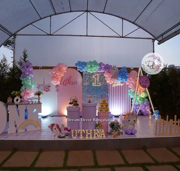 An outdoor first-birthday stage under a white tent: pastel balloon arches, a sequin panel with a lit 1, three round plinths, giant ONE letters, the child's name in lit letters and a picket fence