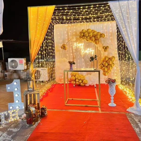 A terrace setup at night: a frame hung with fairy lights and curtains over a red carpet, with a gold and white balloon ring and a lit number 1
