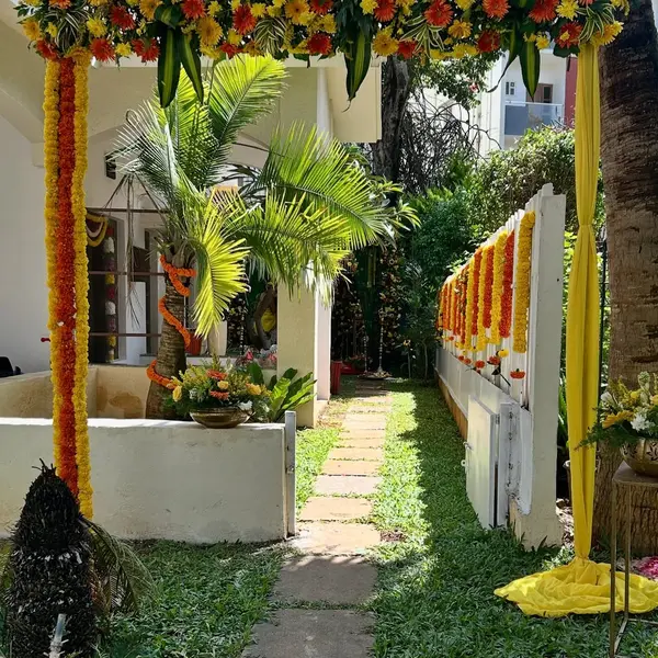 A header of yellow and red gerbera, marigold and palm leaves on two posts wrapped in marigold, with yellow drapes, over a stone path through a garden to a house