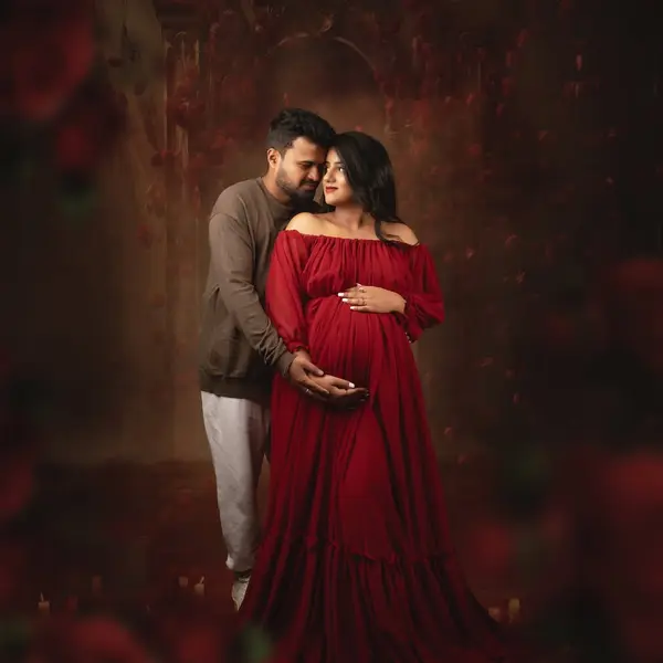 A pregnant woman in a flowing red off-shoulder gown leans into her husband against a dark backdrop of climbing red roses, lit softly from one side