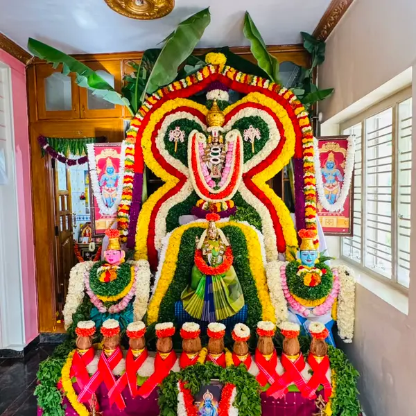 A home pooja in fresh flowers: a tall arch of yellow, red and white garlands behind a goddess idol in a green silk saree, a smaller idol on each side and a row of coconuts tied in red cloth in front