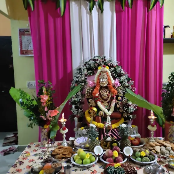 A goddess idol at home in a halo of roses, carnations and baby's breath, with magenta and white drapes under a flower valance, banana leaves, two silver lamps and a table of fruit and offerings
