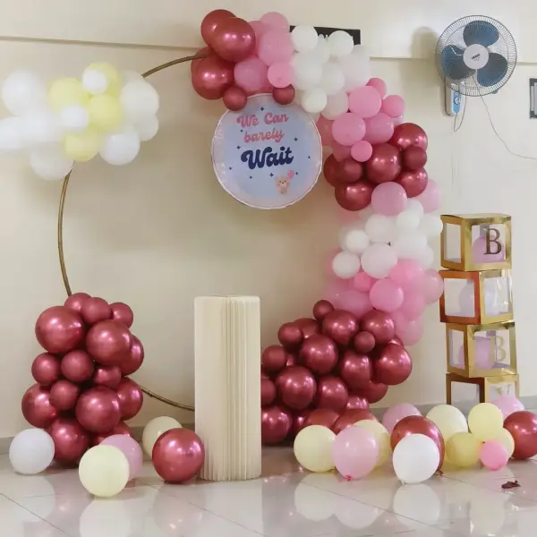 A gold ring with a garland of maroon, pink, white and pale yellow balloons, a round We Can barely Wait sign with a small teddy, a cream pleated plinth and a stack of gold BABY boxes, against a cream wall with a fan