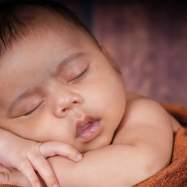 A newborn fast asleep on a rust-brown blanket, both hands folded under the cheek