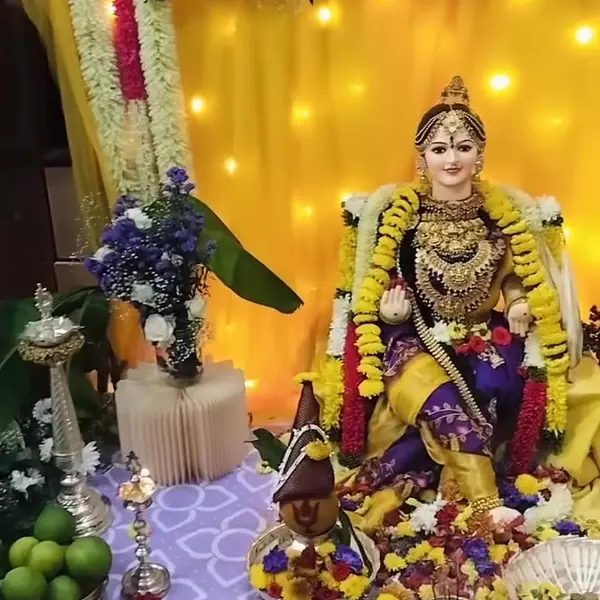 A Varamahalakshmi pooja corner: a golden-yellow drape glowing with fairy lights behind the goddess, white and red flower strings down one side, silver lamps and plates of offerings on a lilac cloth