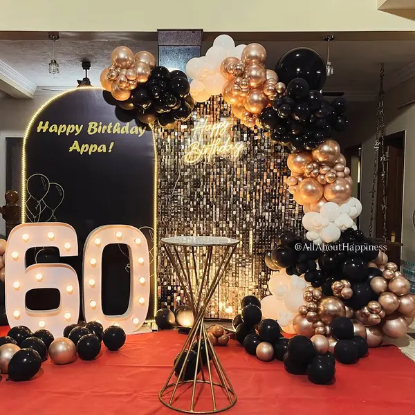 A black arched panel reading Happy Birthday Appa beside a gold sequin wall with a neon sign, a black, rose-gold and white balloon garland, a lit 60 and a tall gold table on a red carpet