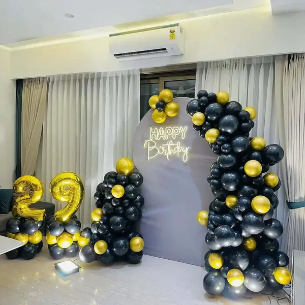 A grey arched backdrop with a warm-white Happy Birthday neon sign, a black and gold balloon garland up one side and gold foil numbers 29 on balloon stands, in a flat with curtains