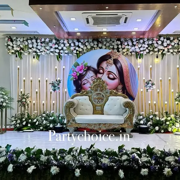 A hall stage: a round print of Yashoda and baby Krishna behind an ornate white and gold sofa, rows of gold candle poles on white drapes, a white flower border on top and a white and purple flower border along the stage front