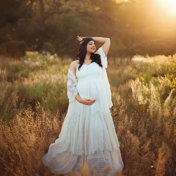 A pregnant woman in a white tiered lace dress and a lace shawl raises one arm in dry grass, the sun low behind the trees
