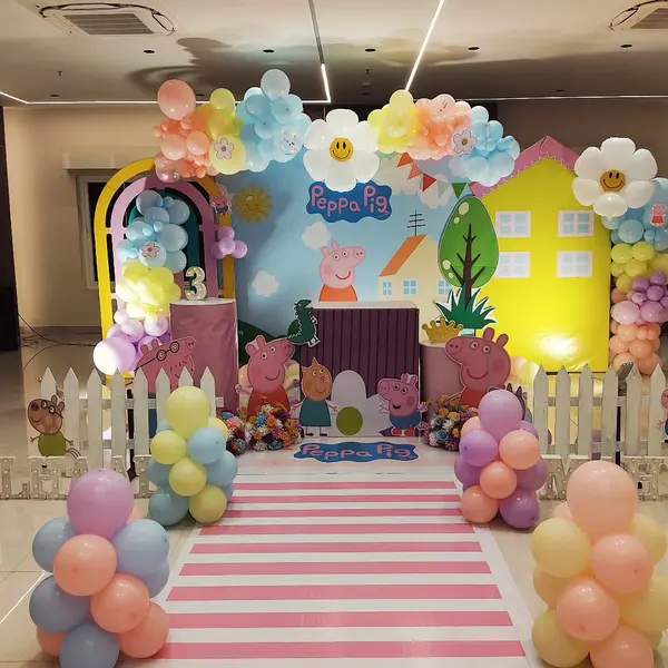 A Peppa Pig stage in an empty hall: a printed backdrop with a yellow house panel, pastel balloon garlands and columns, a white picket fence, character cutouts, plinths, marquee letters along the floor and a pink and white striped runner
