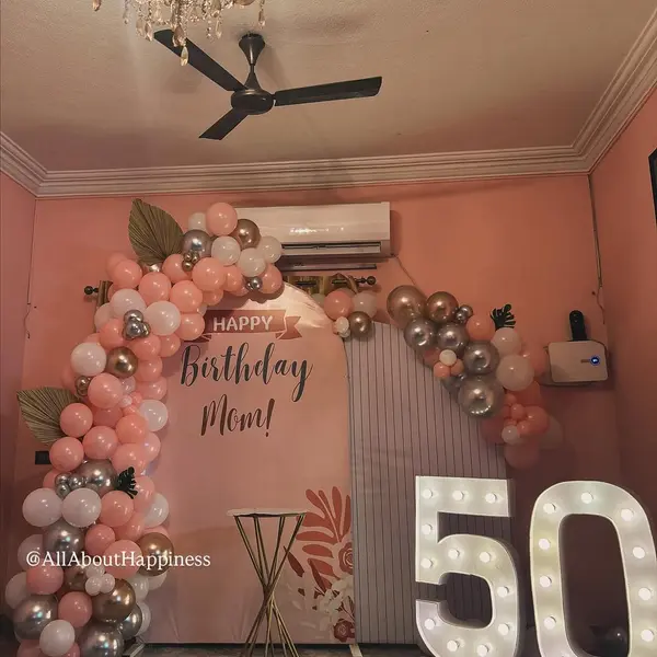 A peach panel reading Happy Birthday Mom with a peach, white and silver balloon arch, a lit 50 and a tall gold table, in a room with peach walls and a ceiling fan