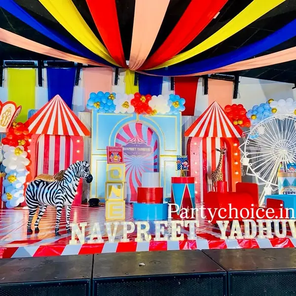 A circus first birthday for twins: ceiling drapes in five colours, two striped tents, a carousel, a Ferris wheel, a life-size zebra, a stage floor printed in red and white and the names in lit letters