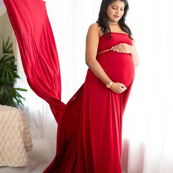 A pregnant woman in a strapless red gown holds her bump while a long red fabric tail flies behind her in a white studio