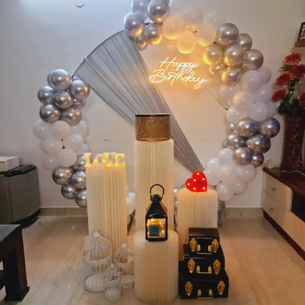 A silver and white balloon ring with a Happy Birthday neon sign and cream plinths, in a living room beside a wooden cabinet
