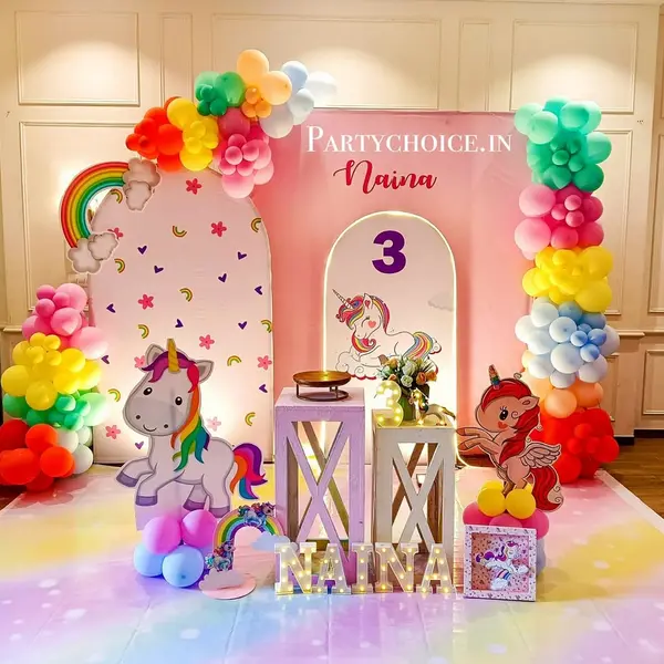 A third birthday: a pink backdrop with two printed arches of hearts, rainbows and a unicorn with a 3, a rainbow garland, two unicorn cutouts, two plinths, the name NAINA in five lit letters and a printed rainbow floor