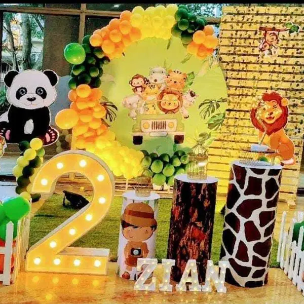 A second birthday: a round jungle print with a jeep and animals in an orange and green garland, tiger, panda, lion, elephant and monkey cutouts, giraffe-print and bark-print plinths, a lit 2, picket fences and the name ZIAN in lit letters