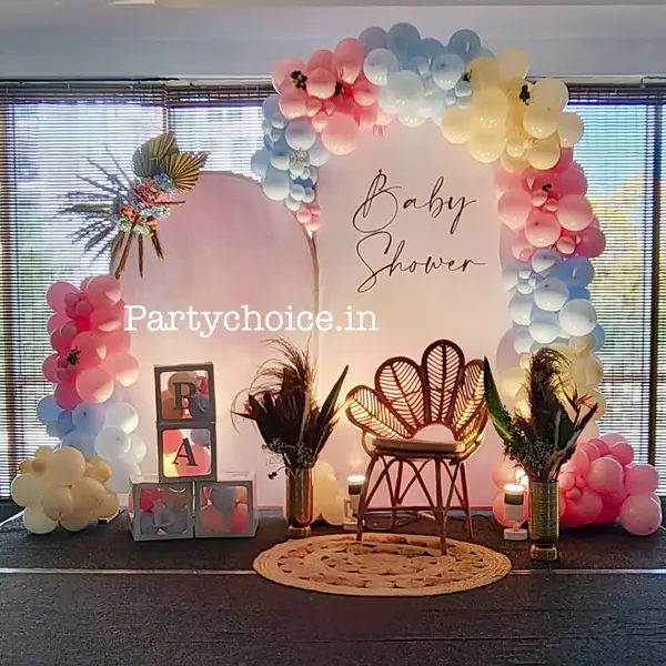 In a hall: a blush arched panel and a white panel reading Baby Shower, a pink, blue, cream and peach balloon garland, dried palm, BABY boxes, a rattan peacock chair, pampas in two gold vases and a round jute rug