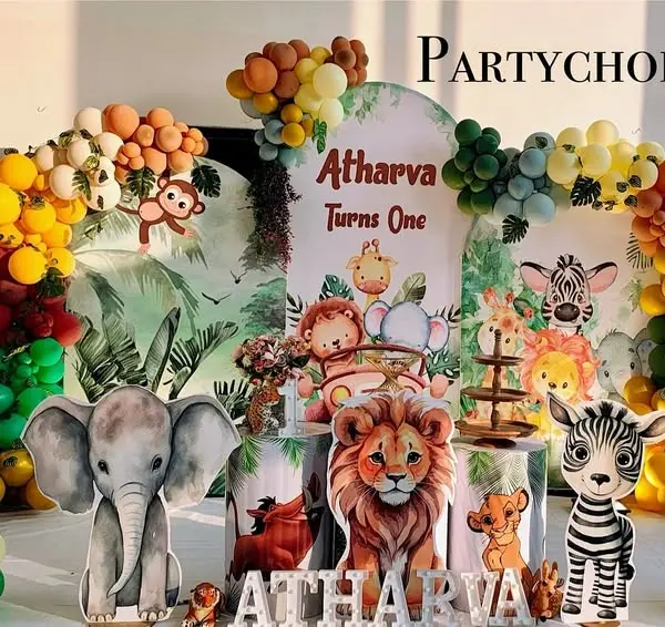A first-birthday stage reading Atharva Turns One: three arched jungle panels, an orange, mustard, red, green and teal garland, tiger, elephant, lion, zebra and life-size giraffe cutouts, printed plinths and the name in lit letters