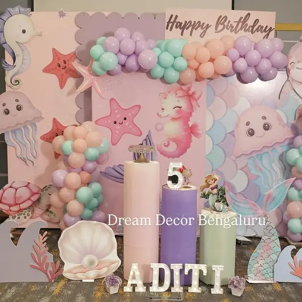 A fifth birthday in a hall: three printed panels with seahorses, jellyfish, starfish and mermaid scales, a teal, lilac and peach garland, seahorse, jellyfish, dolphin and turtle cutouts, three pastel plinths, a lit 5 and the name ADITI in lit letters