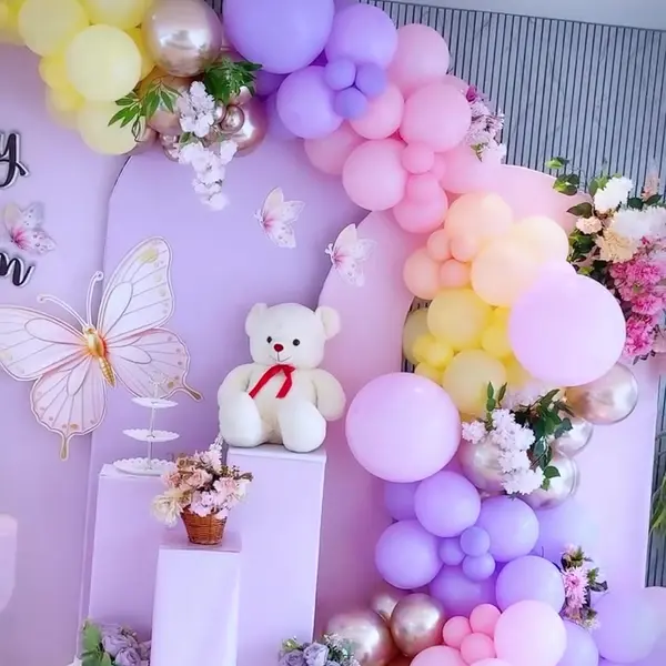 Two lilac U-shaped panels with paper butterflies, a big butterfly cutout, a garland in lilac, pink, yellow and rose gold with flowers, and a white teddy on a lilac plinth, in a hall