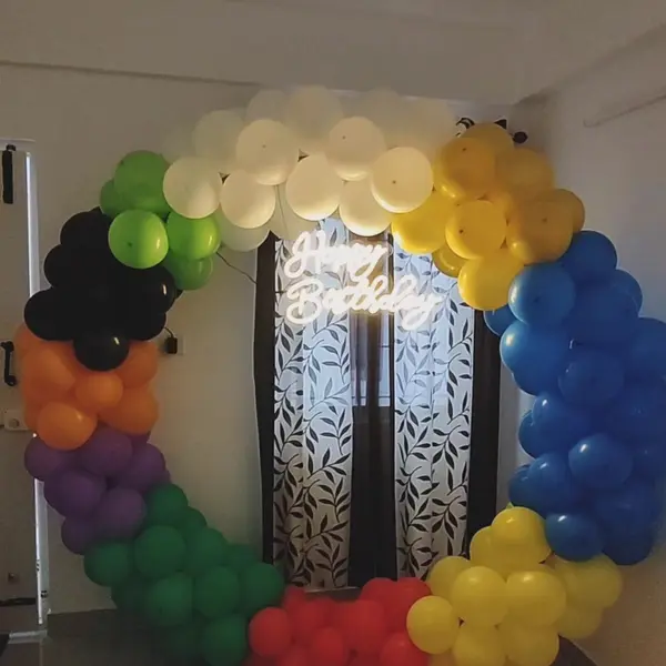 A rainbow balloon ring with a Happy Birthday neon sign, standing in front of a curtained window in a flat