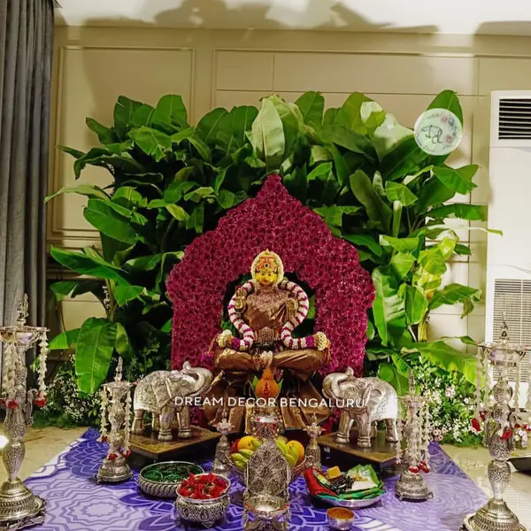 A seated Lakshmi inside a pointed arch of magenta roses, in front of a wall of large green leaves, between two silver elephants and four tall silver lamps, on a purple patterned floor cloth with silver bowls of offerings