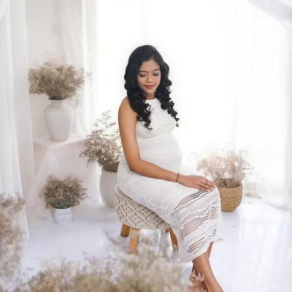 A pregnant woman in a white knit dress sits on a woven stool in a bright white studio set with dried flowers and sheer curtains