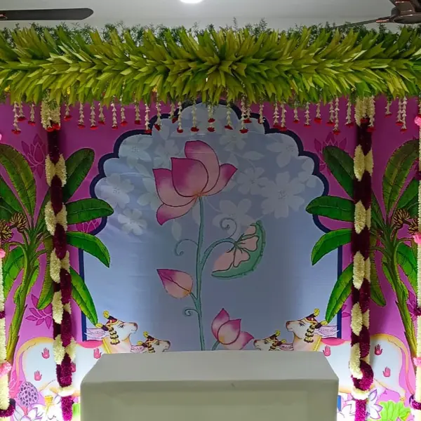 A printed panel of a pink lotus in a scalloped arch, with painted banana plants and cows, strings of white and maroon flowers down both sides, a header of green leaves and a plain white table in front