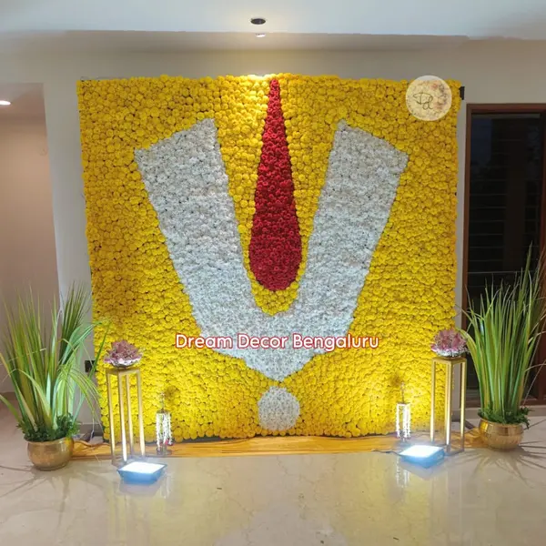 A wall of yellow flowers about 8 feet square with a large namam in white and red flowers, two grass pots and two lights on stands, for a Satyanarayana pooja