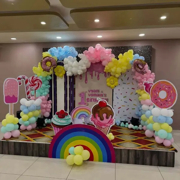 A twins' first birthday on a hall stage: a drip-icing panel and a sprinkle panel, a pastel garland with donut foils, life-size popsicle, candy-cane, lollipop, cupcake, ice-cream and donut cutouts and a big rainbow at the front
