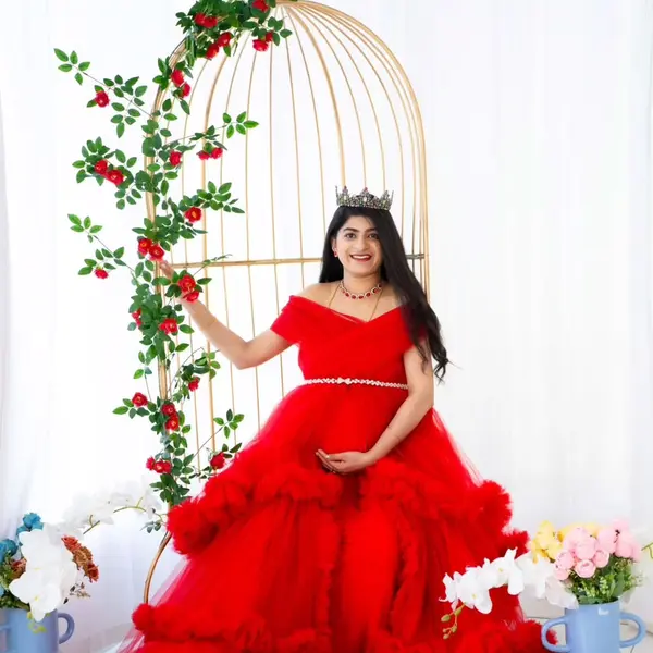 A pregnant woman in a red ruffled tulle gown with a pearl belt and a crown sits by a tall golden birdcage wound with a red rose vine, white flowers at her feet