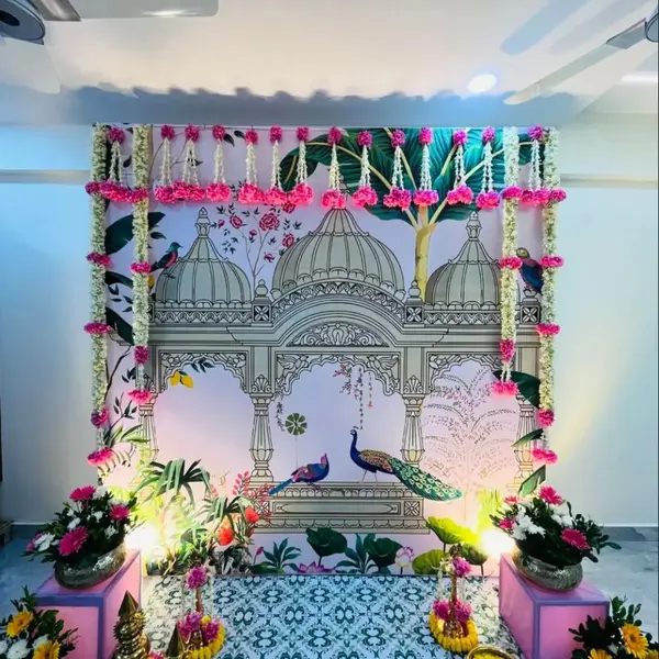 A printed Pichwai backdrop of domes and peacocks at a housewarming, with pink and white flower strings across the top and down the sides, flower pots on pink stands and brass lamps on rings of petals