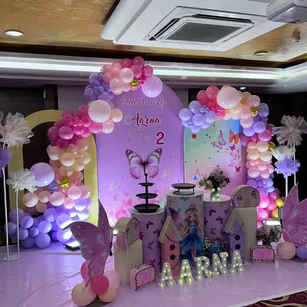 A butterfly second-birthday stage in a hotel hall: layered purple and pastel backdrops washed in coloured light, two balloon arches, butterfly cutouts, pleated plinths and the name AARNA in lit letters on a lilac floor