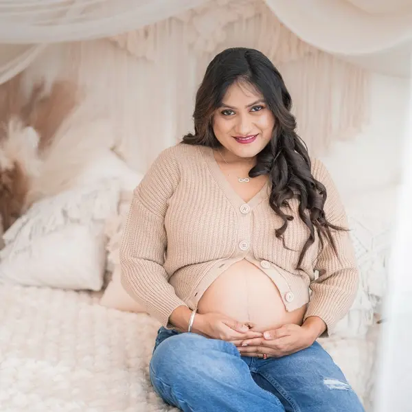 A pregnant woman in a cream cardigan and blue jeans sits on a white woven throw with her hands on her bare bump, white drapes, macramé and pampas behind