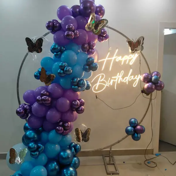 A ring stand with a purple, blue and chrome balloon garland down one side, gold and silver butterflies, and a warm Happy Birthday neon sign inside the ring, against a plain wall