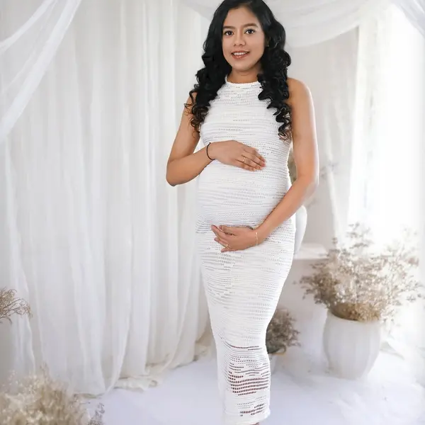 A pregnant woman in a long white crochet dress stands against white drapes, with dried flowers in pots, a stool and a white floor