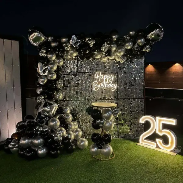 A silver sequin wall with a Happy Birthday neon sign, a black and silver balloon garland over it, a small round table and a lit 25, on artificial grass at night