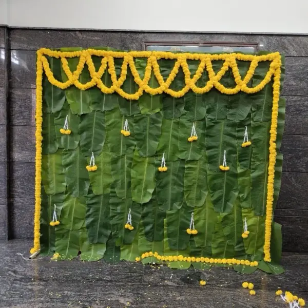 A wall of fresh banana leaves with a marigold swag across the top, marigold strings down both sides and small hanging marigold drops, against a dark granite wall